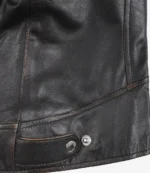 Close-up of a black leather jacket's lower section, featuring a silver snap button and a decorative strap. The leather appears smooth and slightly worn.